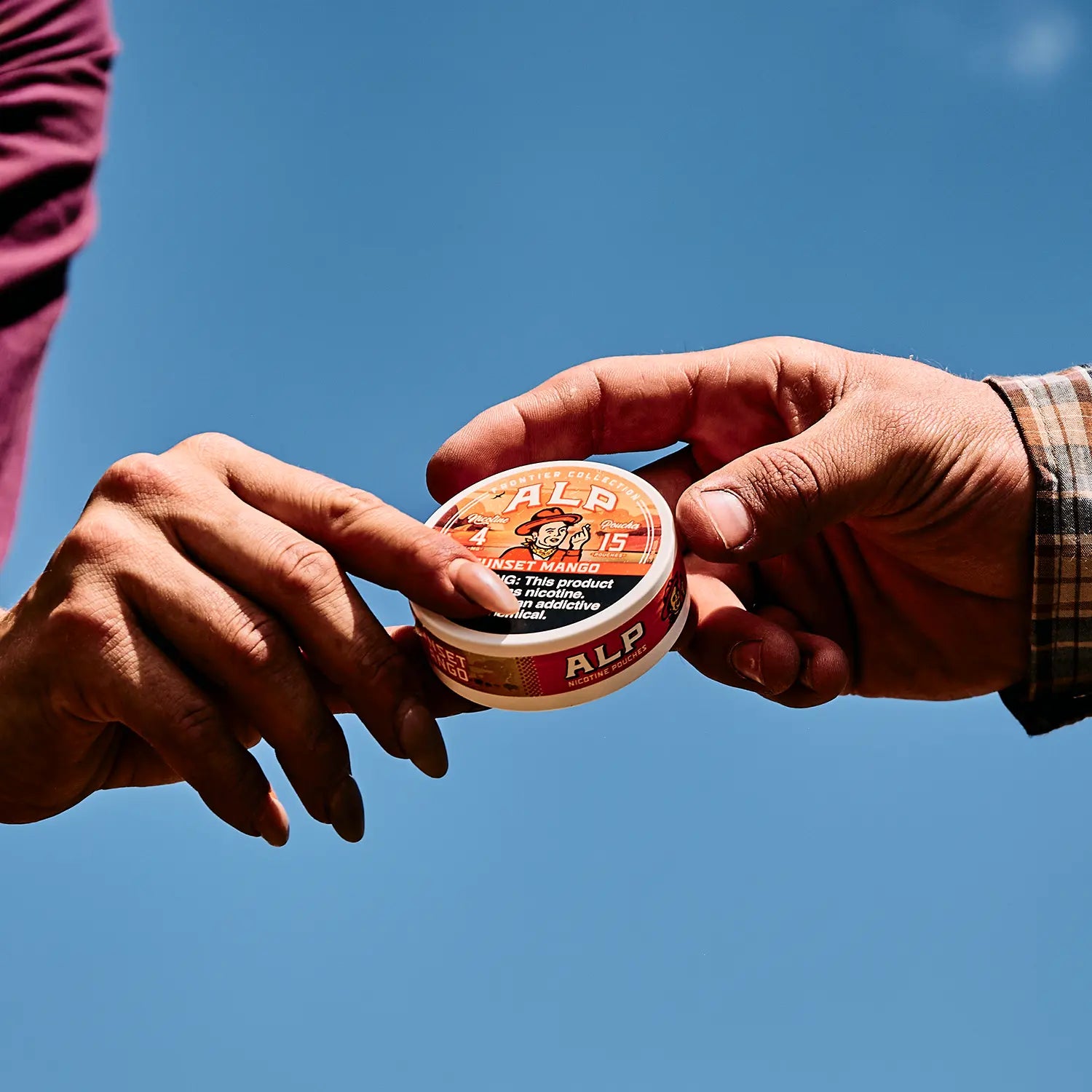 Hand passing an ALP Frontier Collection sunset mango nicotine pouches tin against a clear blue sky
