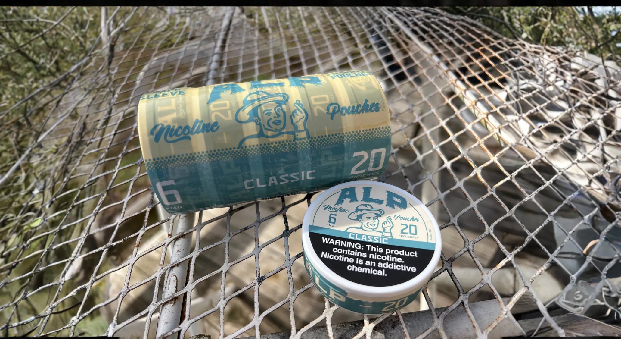 ALP Classic 6mg modern nicotine consumption multi-pack sleeve and single tin resting on a wire fence with greenery visible in the background.