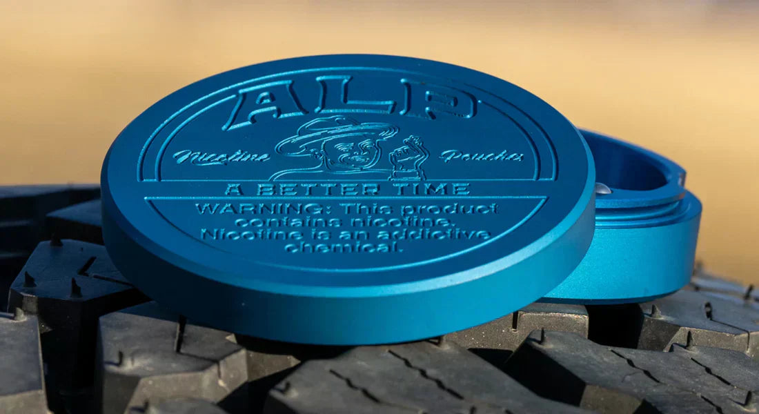 Oral nicotine product displayed as a blue ALP metal tin resting on a rugged tire surface for outdoor appeal.
