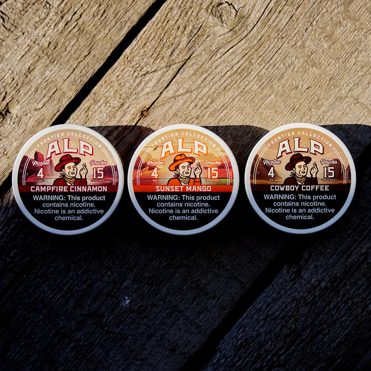 Three ALP Frontier Collection nicotine pouches tins on a rustic wood surface, showing Campfire Cinnamon, Sunset Mango, and Cowboy Coffee flavors